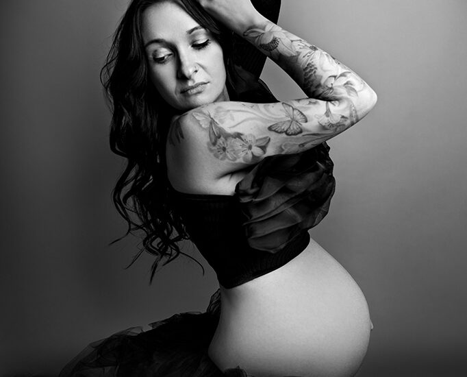 Foto Fine Art in bianco e nero di maternità in studio, di una donna incinta tatuata con un velo nero sul bacino, mentre guarda verso il basso. Fotografia in bianco e nero di maternità in studio con donna tatuata Trieste