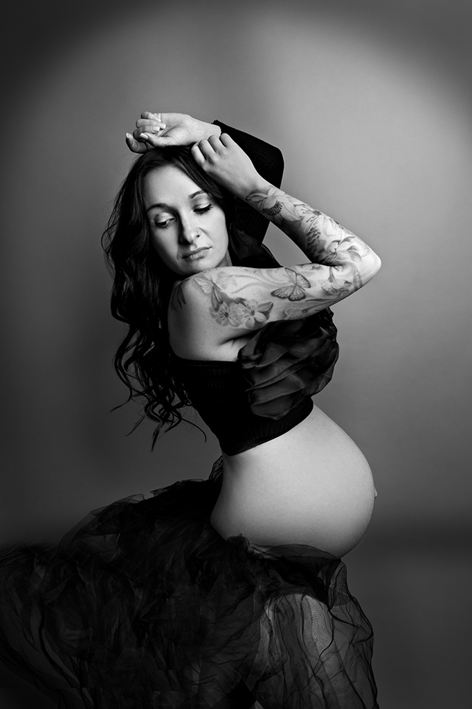 Foto Fine Art in bianco e nero di maternità in studio, di una donna incinta tatuata con un velo nero sul bacino, mentre guarda verso il basso. Fotografia in bianco e nero di maternità in studio con donna tatuata Trieste