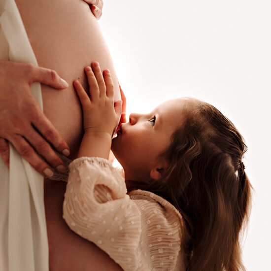 Foto a colori di maternità in studio, di una donna incinta, mentre la bambina le bacia il pancione. Fotografia a colori di maternità con bambina in studio Italia