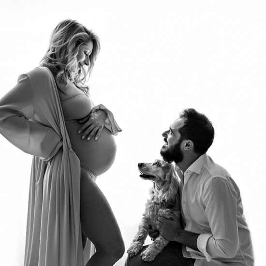 Foto in bianco e nero di famiglia, di una donna incinta, con il compagno ed il cane che la guardano. Fotografia di maternità di famiglia in bianco e nero Italia