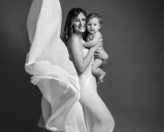 Foto ritrattistica in bianco e nero di una mammacon un vestito bianco di tulle svolazzante dietro di lei ed il bambino nudo in braccio. Fotografia ritrattistica in bianco e nero Trieste
