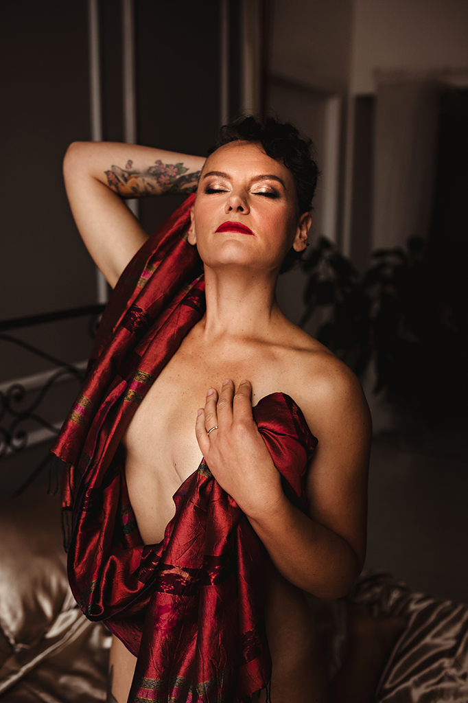 Foto a colori di una donna nuda con un velo rosso sul petto, in ginocchio sul letto. Fotografia donna sensuale boudoir Italia