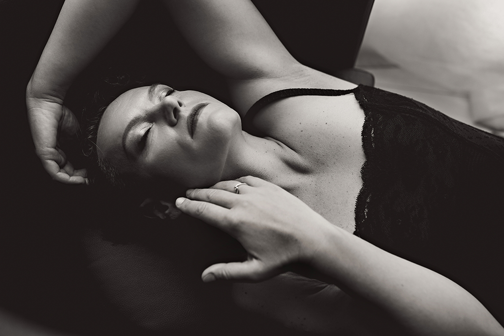 Foto in bianco e nero di una donna con una canotta nera di pizzo, distesa con le mani sul viso. Fotografia sensuale donna boudoir Trieste