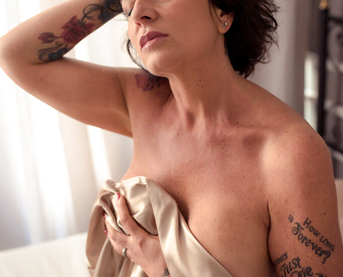 Foto a colori di una donna tatuata nuda, su un letto, mentre si copre con un velo in raso beige. Fotografia a colori donna sensuale boudoir Italia