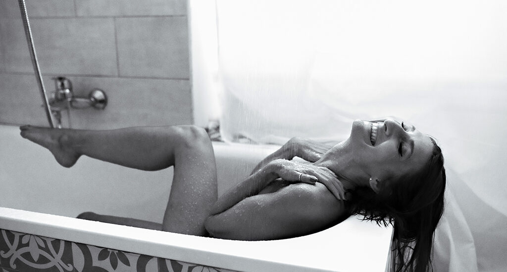 Foto in bianco e nero di una donna nuda nella doccia con le mani sulle spalle, con l'acqua che scorre addosso, mentre sorride. Fotografia donna boudoir Trieste