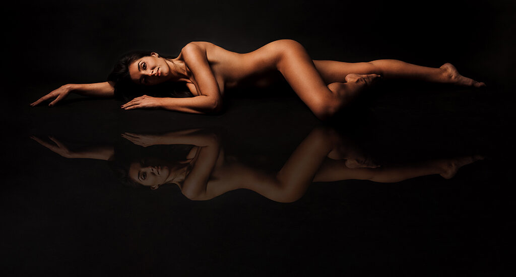Foto a colori artistica di una donna nuda distesa per terra, con il riflesso. Fotografia sensuale donna nuda distesa per terra boudoir Italia