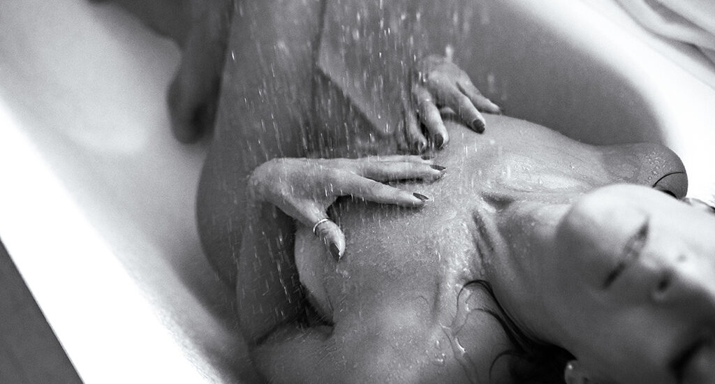 Foto in bianco e nero di una donna nuda nella doccia con le mani sul petto, con l'acqua che scorre addosso. Fotografia donna boudoir Trieste