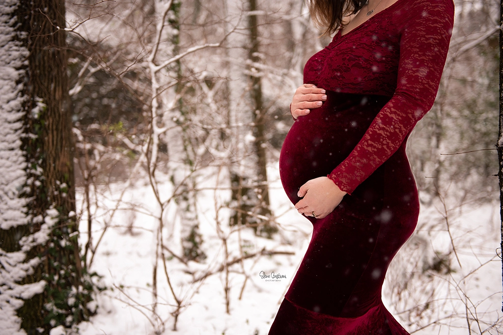 Fotografia a colori fuori studio di una donna incinta con un vestito rosso scuro, risaltando la pancia con le mani della donna sopra. Fotografia a colori di una donna incinta sulla neve Trieste