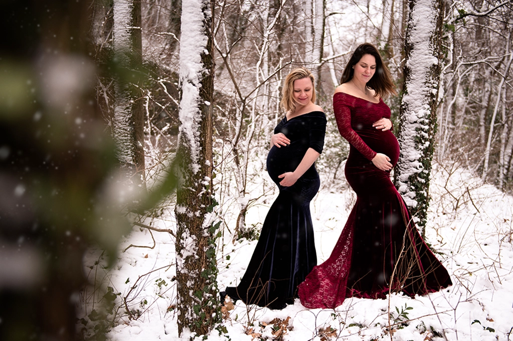 Foto a colori fuori studio di due donne incinte in un bosco con la neve, mentre entrambe si tengono la pancia. Fotografia a colori fuori studio di maternità con due donne incinte Italia