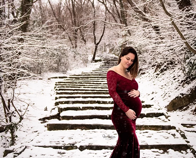 Foto a colori di una donna incinta sulla neve con un vestito rosso scuro, mentre si tiene la pancia con lo sguardo verso il basso. Fotografia a colori di maternità fuori studio sulla neve Trieste