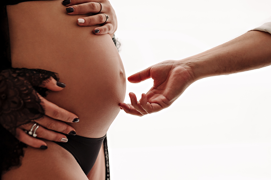 Foto a colori di maternità in studio, di una donna incinta con la mano del compagno che sfiora la pancia della donna. Fotografia a colori di maternità in studio Italia