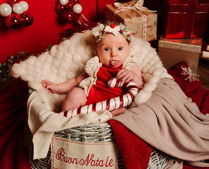 Foto a tema natalizio di una bimba in un cesto tra le coperte, che tiene in mano un bastonicino di zucchero. Fotografia di una bimba in un cesto a tema natalizio Italia