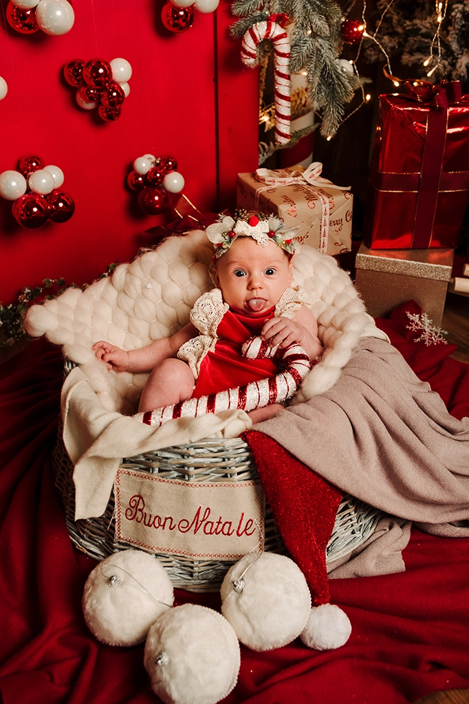 Foto a tema natalizio di una bimba in un cesto tra le coperte, che tiene in mano un bastonicino di zucchero. Fotografia di una bimba in un cesto a tema natalizio Italia