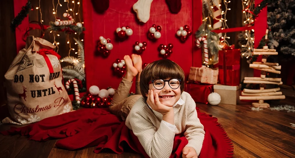Foto a tema natalizio di un bambino disteso per terra con una mano sul viso, sopra ad una coperta rossa. Fotografia di un bambino a tema natalizio Italia