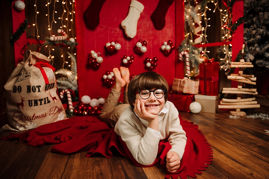 Foto a tema natalizio di un bambino disteso per terra con una mano sul viso, sopra ad una coperta rossa. Fotografia di un bambino a tema natalizio Italia