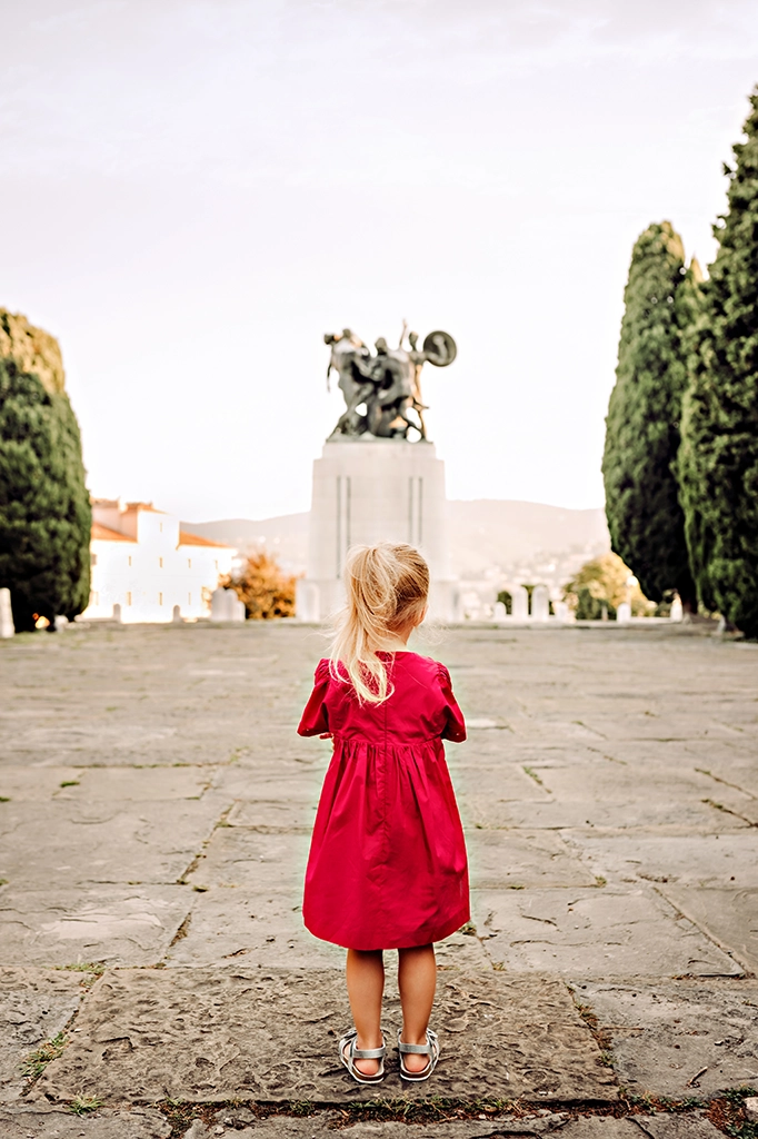 Foto a colori fuori studio di una bambina con un vestito rosso, messa di schiena mentre guarda la statua di fronte. Fotografia fuori studio a colori di una bambina Italia