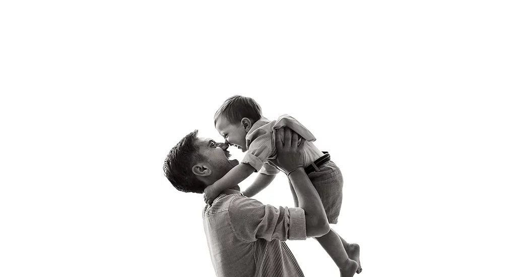 Foto in bianco e nero in studio di famiglia, mentre il papà tiene in braccio il bambino e si guardano ridendo, naso contro naso. Fotografia in bianco e nero di famiglia papà e bambino Trieste