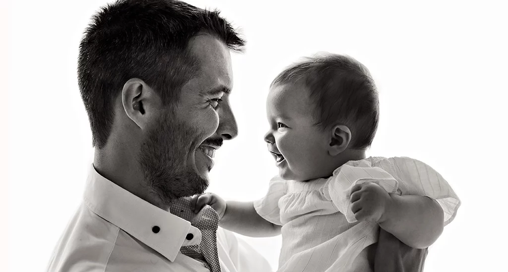 Foto in bianco e nero in studio di famiglia, mentre il papà tiene in braccio il bambino e si guardano ridendo. Fotografia in bianco e nero di famiglia papà e bambina Trieste