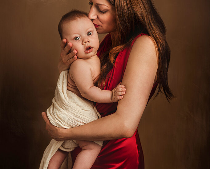 Foto a colori in studio di famiglia, mentre la mamma, con un vestito rosso, tiene in braccio il bambino baciandolo sulla testa. Fotografia a colori di famiglia mamma e bambino Italia