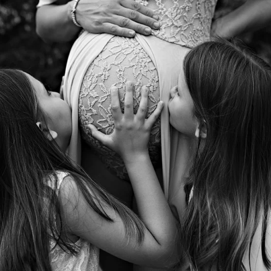 Foto in bianco e nero fuori studio di una donna incinta mentre due bambine baciano il pancione. Fotografia in bianco e nero fuori studio maternità Trieste