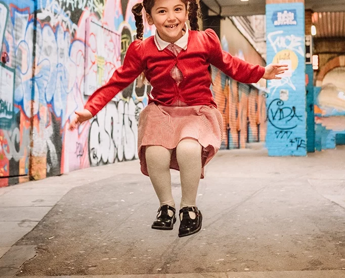 Foto a colori fuori studio di una bambina con le trecce, mentre salta, vestita di rosso. Fotografia a colori di una bambina che salta Trieste