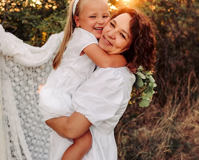 Foto a colori fuori studio di una mamma con in braccio la figlia, entrambe vestite di bianco, mentre si abbracciano sorridendo. Fotografia a colori di una mamma con la figlia in braccio Trieste