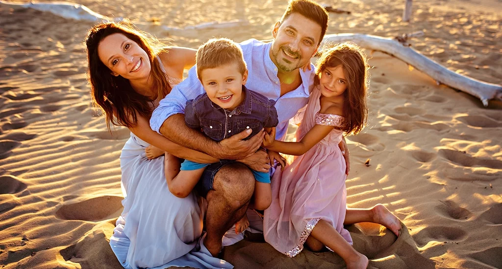 Foto a colori fuori studio di famiglia seduti sulla spiaggia al tramontare del sole in cui tutti guardano nell'obiettivo sorridendo. Fotografia fuori studio di famiglia Trieste