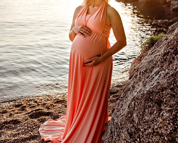 Foto a colori di maternità in riva al mare, di una donna incinta con un vestito lungo color salmone, mentre si tiene la pancia. Fotografia a colori di maternità in riva al mare Italia