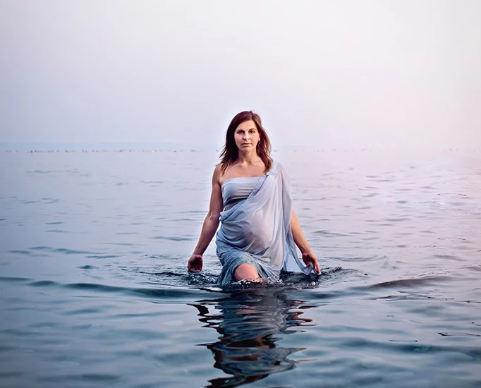 Foto a colori fuori studio nel mare di una donna incinta con un vestito azzurro, mentre cammina. Fotografia a colori di maternità sul mare Italia