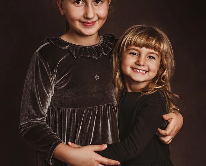 Foto ritratto a colori di due sorelle che si abbracciano ed entrambe sorridono. Fotografia a colori ritrattistica Trieste