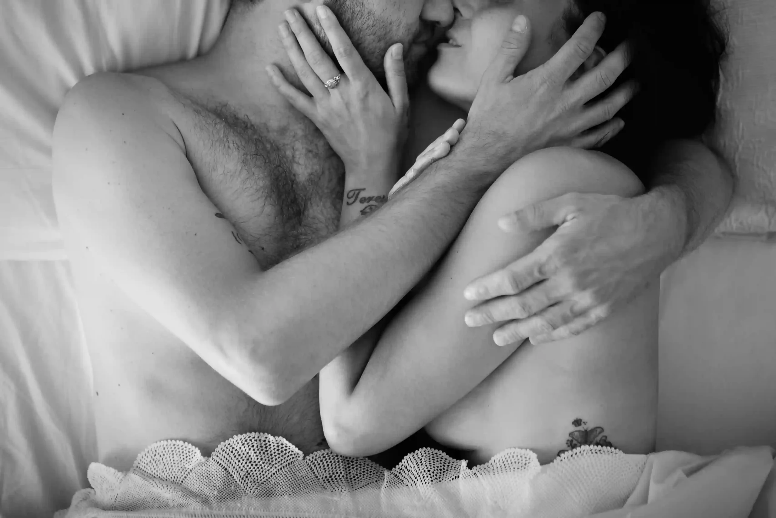 Coppia abbraccciata in un momento intimo a letto, posti entrambi lateralmente con le mani sul viso dell'altro, sotto una coperta con il pizzo. Fotografia di coppia boudoir bianco e nero Italia