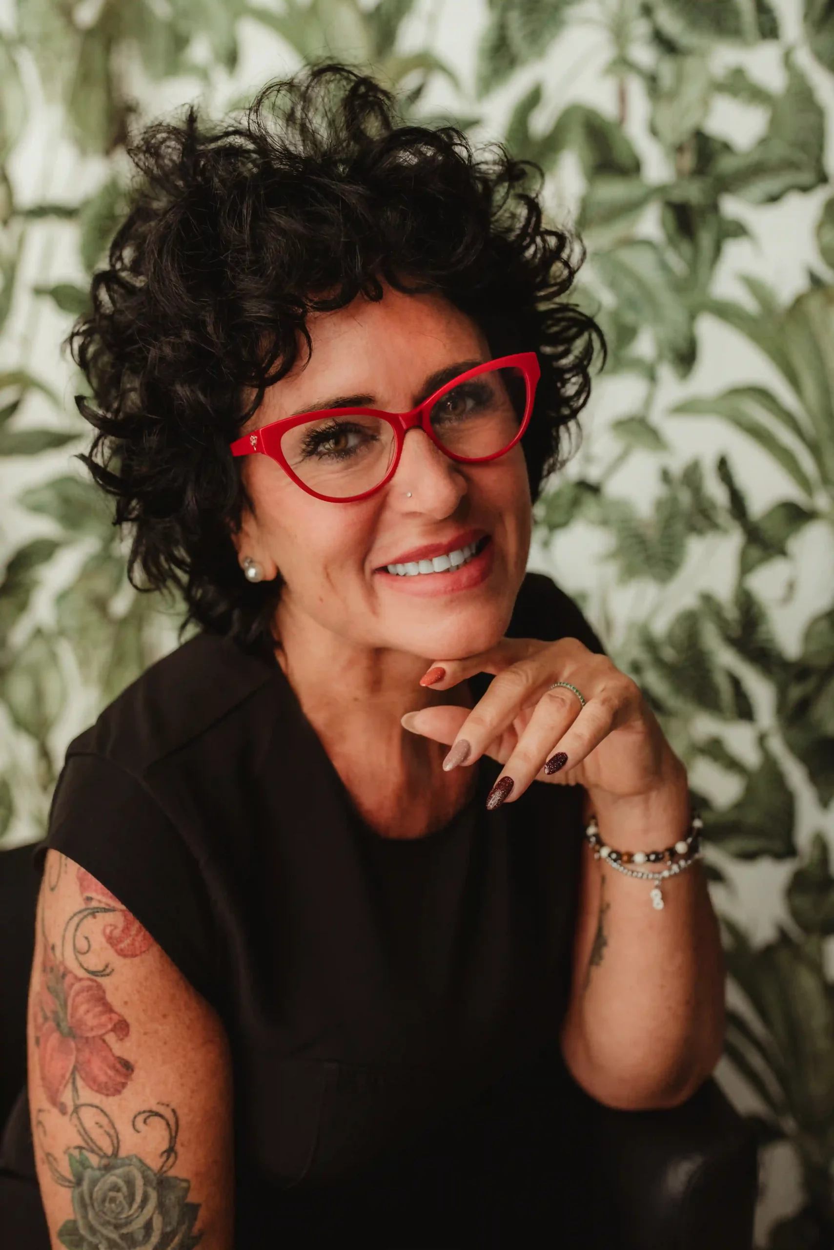 Foto ritratto business a colori donna tatuata che sorride con occhiali rossi e tiene la mano sotton il viso. Fotografia ritratto a colori Italia