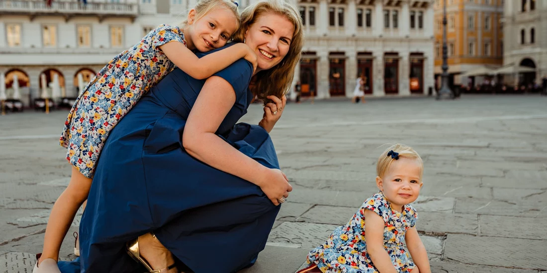 Foto a colori fuori studio, di una famiglia in cui la bambina abbraccia la madre sorridendo, con l'altra bambina seduta per terra di fronte a loro. Fotografia a colori di famiglia Italia