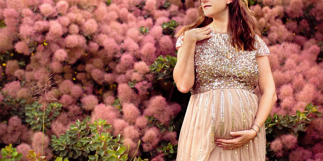 Foto a colori fuori studio di una donna incinta con un vestito lungo rosa con i brillantini, davanti ad un cespuglio di fiori rosa. Fotografia fuori studio a colori di maternità Trieste
