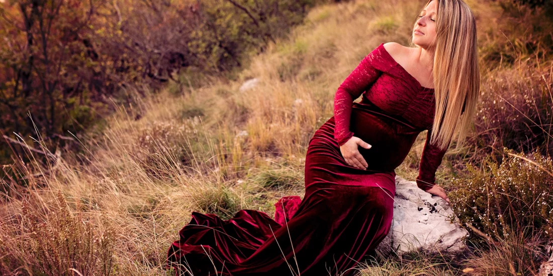 Foto a colori di una donna bionda incinta con un vestito rosso scuro di velluto, mentre si tiene la pancia. Fotografia a colori di maternità fuori studio Trieste